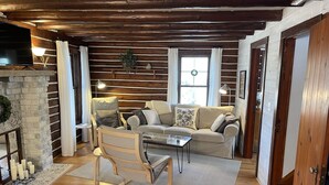 A cozy living room with a stone fireplace, wooden beams, a sofa, a coffee table, and a TV. - Updated Vintage 1938 Log Cabin – Walk to Sister Bay (Sister Bay)
