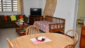 A living room with a wooden dining table, chairs, a sofa, a television, and a patterned bedspread. - Aito1 across from Muri Beach (Muri Beach)