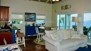 A spacious living area with a white sofa, a dining table with chairs, a kitchen area, and a view of the ocean through large windows.