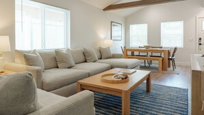 A modern living room with a sectional sofa, a wooden coffee table, and a dining area with a table and chairs. - Beach Please ~ Luxe Cottage ~ Steps from Sand (Treasure Island)