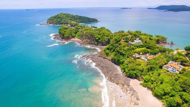 An aerial view of a coastal area with lush greenery, a beach, and several buildings. - Flamingo Estates 18- 4 BR House with Pool (Flamingo)