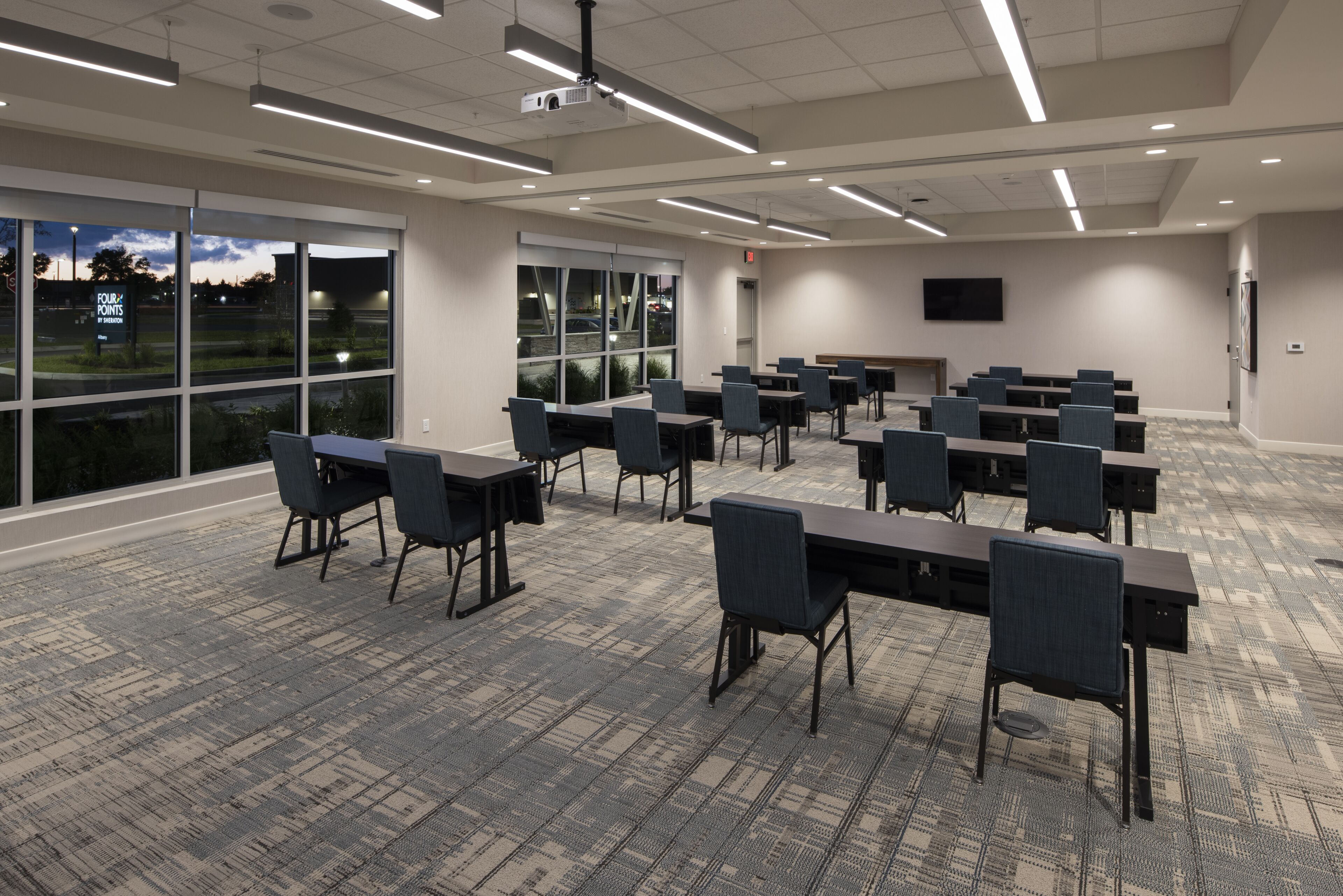 Une salle de conférence équipée de tables et de chaises disposées pour les réunions, de grandes fenêtres offrant une vue sur l’extérieur et d’un téléviseur à écran plat fixé au mur.