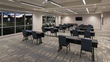 Une salle de conférence équipée de tables et de chaises disposées pour les réunions, de grandes fenêtres offrant une vue sur l’extérieur et d’un téléviseur à écran plat fixé au mur.