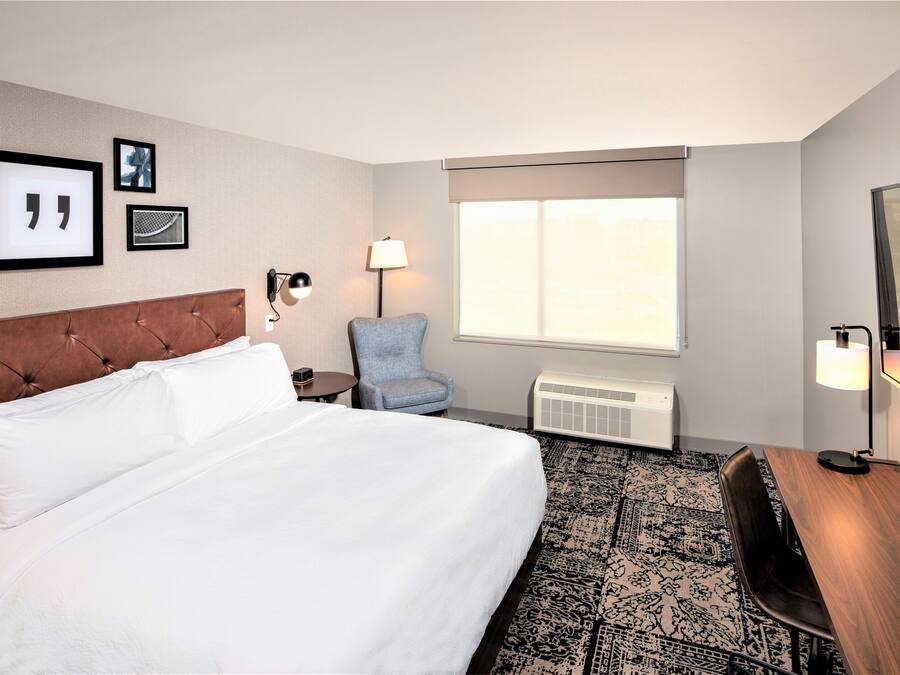 Quarto de hotel com uma cama grande, uma mesa, uma cadeira, uma janela com vista e luminárias fixadas na parede.
