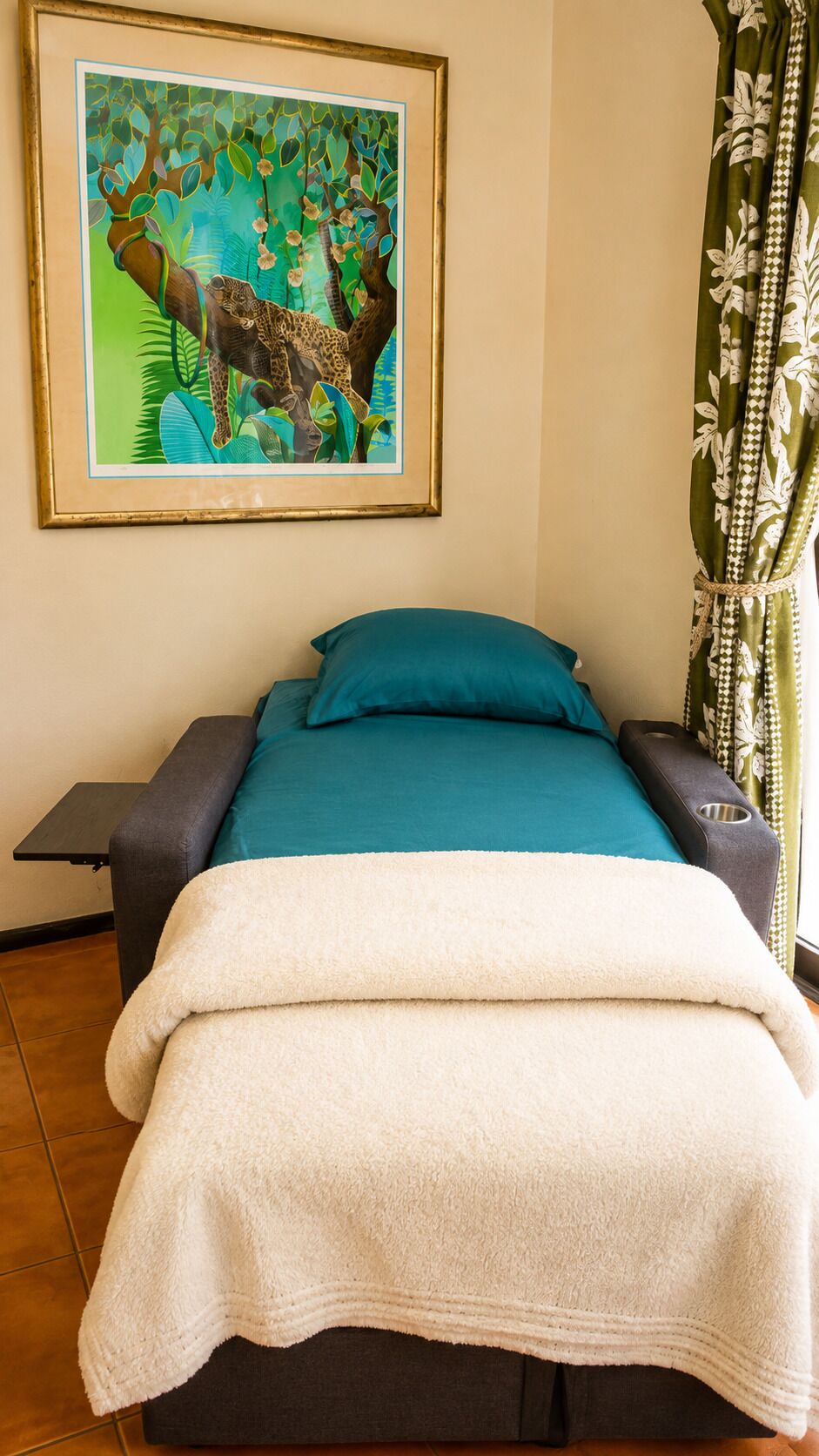 A bed with a teal bedspread and a white blanket, a framed painting of a jungle scene on the wall, and a curtain with a leaf pattern.