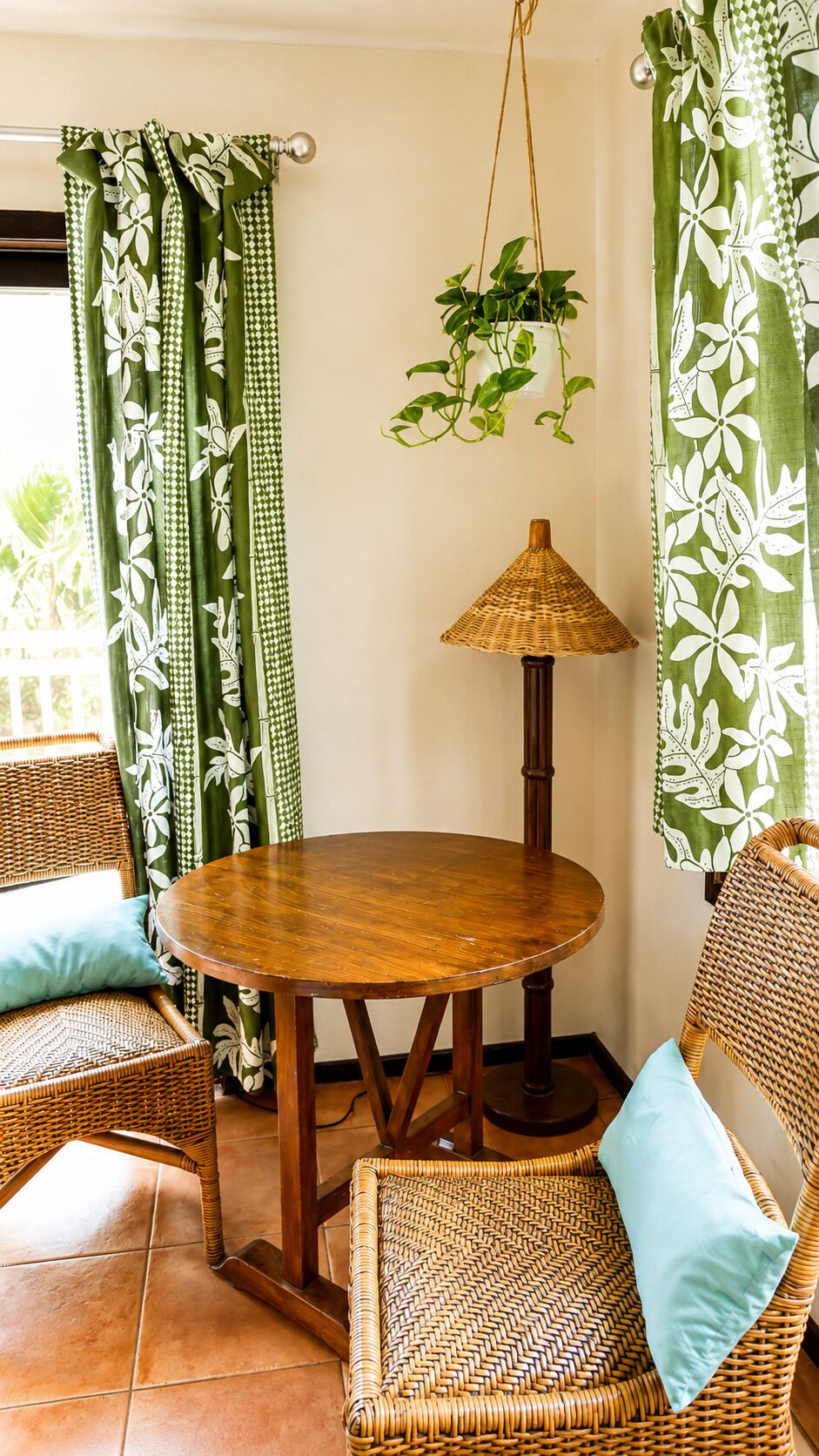 A room with a round wooden table, two wicker chairs, a hanging plant, and green patterned curtains.