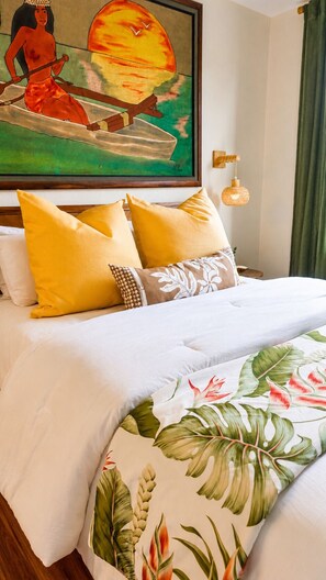 Une chambre d’hôtel avec un lit, des oreillers, une couverture aux motifs tropicaux, un tableau représentant un coucher de soleil et une lampe fixée au mur.