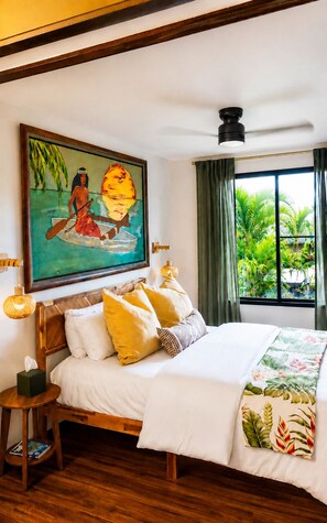 Une chambre d’hôtel avec un lit, un tableau représentant un paysage tropical, une fenêtre avec des rideaux et une vue sur la verdure à l’extérieur.