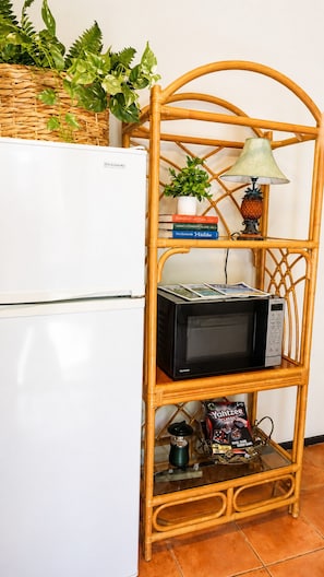 A wicker shelf unit with a microwave, books, a small plant, and a bottle of Yachtzee Classic beer. - An Affordable Alternative to Expensive Accommodations in Poipu (Poipu)