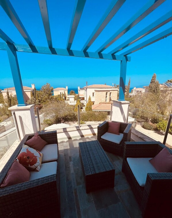 A covered outdoor seating area with wicker furniture and cushions, overlooking a coastal town.
