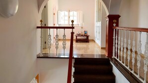 A grand staircase with ornate white balustrades leading to a spacious room with a large window and a small table with a lamp. - Happy Family Villa (Arucas)