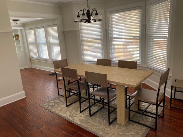 A dining room with a wooden table and chairs, large windows with blinds, and a chandelier.