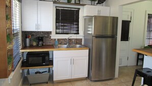 A compact kitchen with white cabinets, a stainless steel refrigerator, a microwave, and a window with blinds. - The Hideaway (Great Bend)