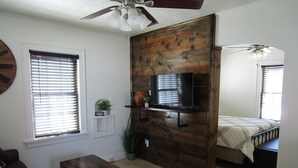 A living room with a wooden accent wall, a television mounted on it, a sofa, a coffee table, and a bed with a striped bedspread. - The Hideaway (Great Bend)