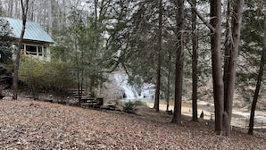 A rustic cabin with a metal roof, surrounded by a forested area with a waterfall in the background. - Waterfall Cottage within sight & sound of Cane Creek Falls, 2BR 2BA (Dahlonega)