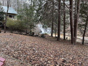 A rustic cabin with a metal roof, surrounded by a forested area with a waterfall in the background.