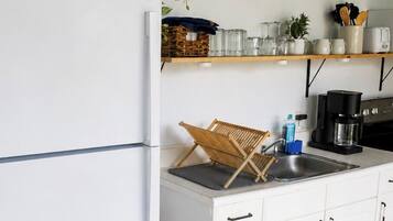 Una cocina con un refrigerador blanco, un lavamanos y una cafetera. Hay un estante sobre el lavamanos con cristalería y una planta.