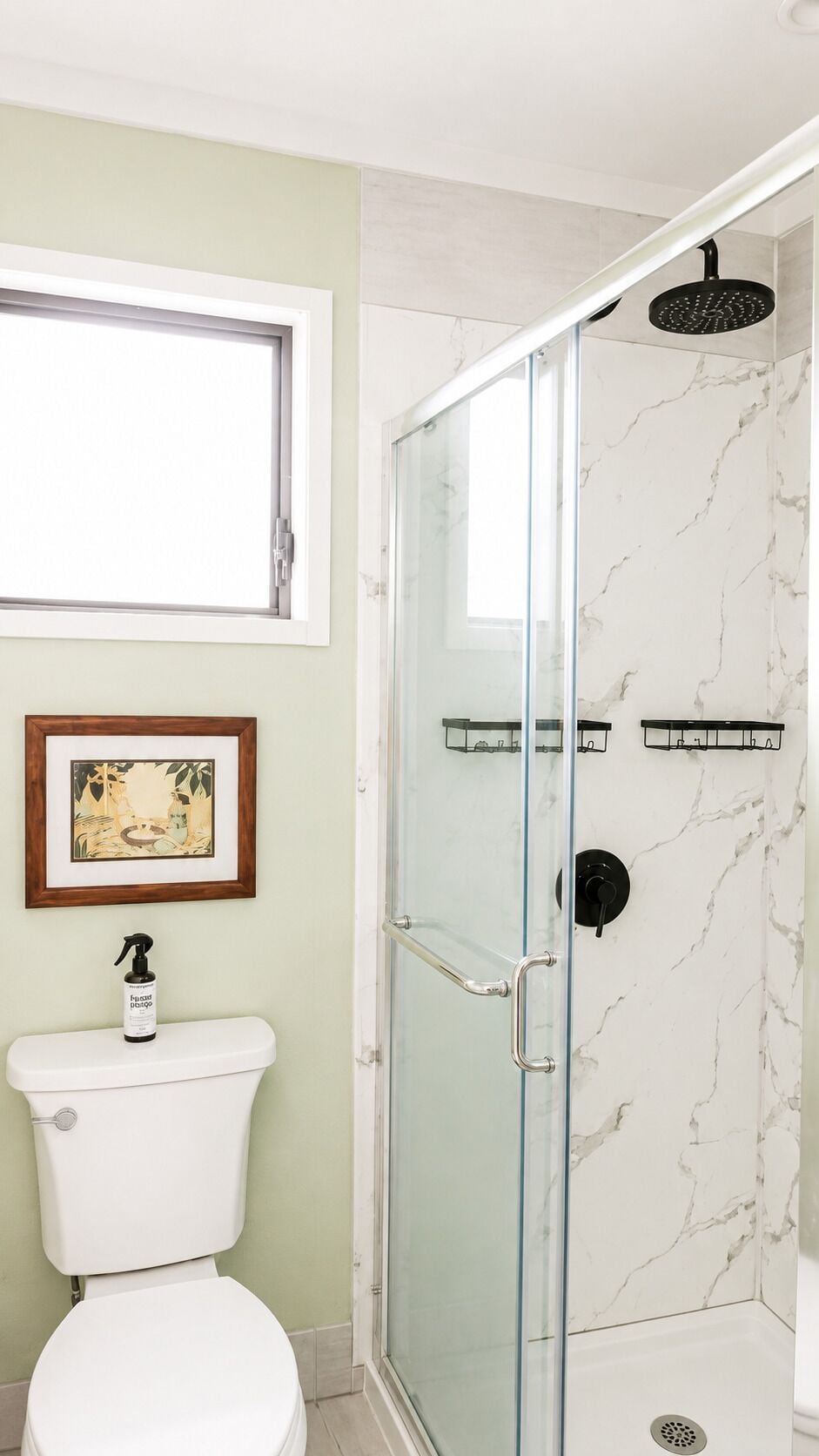 A bathroom with a glass-enclosed shower, a toilet, a framed picture on the wall, and a bottle of shampoo.