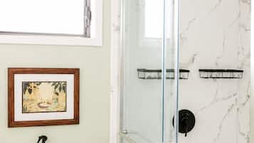 A bathroom with a glass-enclosed shower, a toilet, a framed picture on the wall, and a bottle of shampoo.
