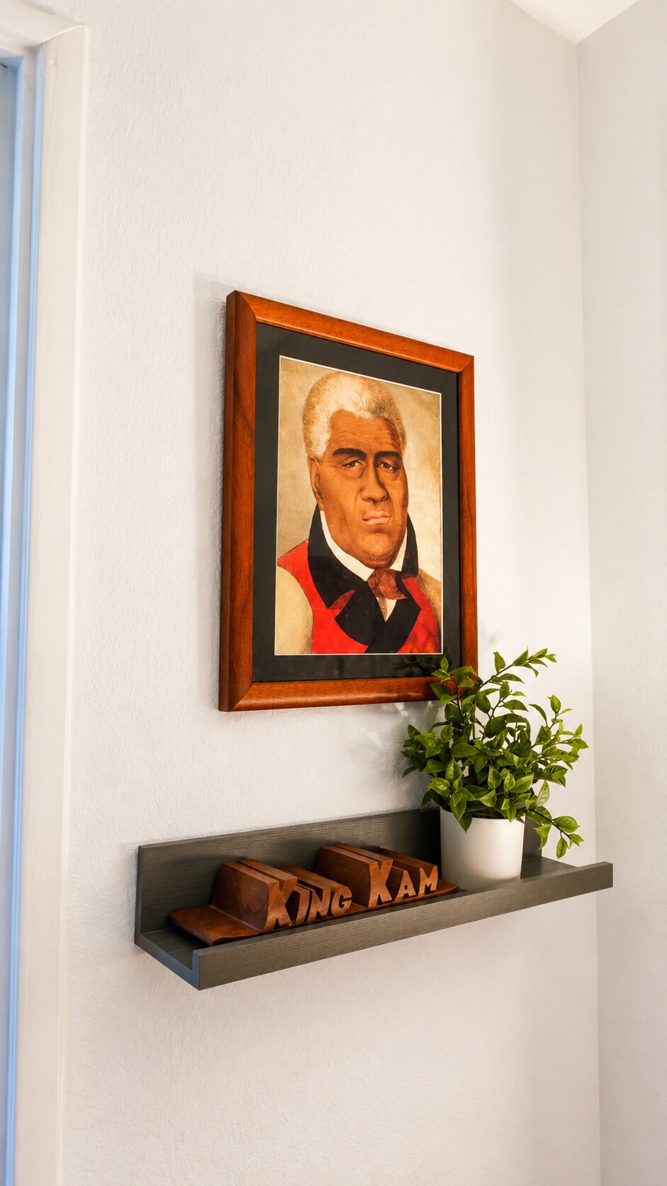 Un retrato en un marco colgado en una pared, un estante con una planta en maceta y un bloque de madera con el texto 'KING KAM'.
