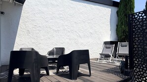 An outdoor patio with a wooden deck, two black plastic chairs, a small table, and a grill. - MAISON LITTLE WHITE HOUSE (Anglet)