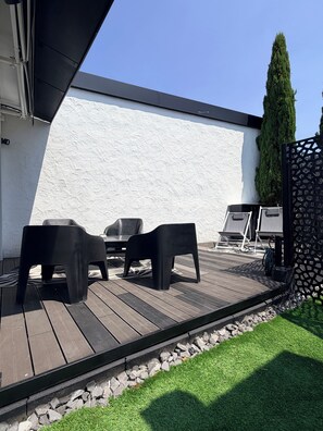 An outdoor patio with a wooden deck, two black plastic chairs, a small table, and a grill.