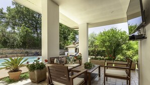 A covered outdoor seating area with a pool, potted plants, and a stone wall. - La Villa - Luxury Home (Configni)
