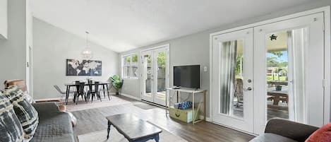 Amplia sala de estar con sofá, mesa de centro y televisor. Se observa un comedor con mesa y sillas, y un patio con mesa y sillas visibles a través de puertas de vidrio.