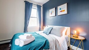 Habitación de hotel con cama, ventana con cortinas, mesita de noche con lámpara y dos cuadros en la pared.