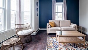 Un salón con una pared de acento azul oscuro, un sofá color beige con un cojín marrón, una silla de mimbre con un cojín blanco, una mesa de centro de madera y una alfombra estampada.