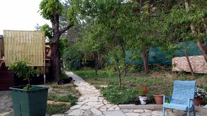 A stone pathway leading to a blue chair and a green trash bin in a garden area with trees and potted plants. - 10 minutes walk from the waterfall, bright apartment, pets welcome! (Saint-Laurent-le-Minier)