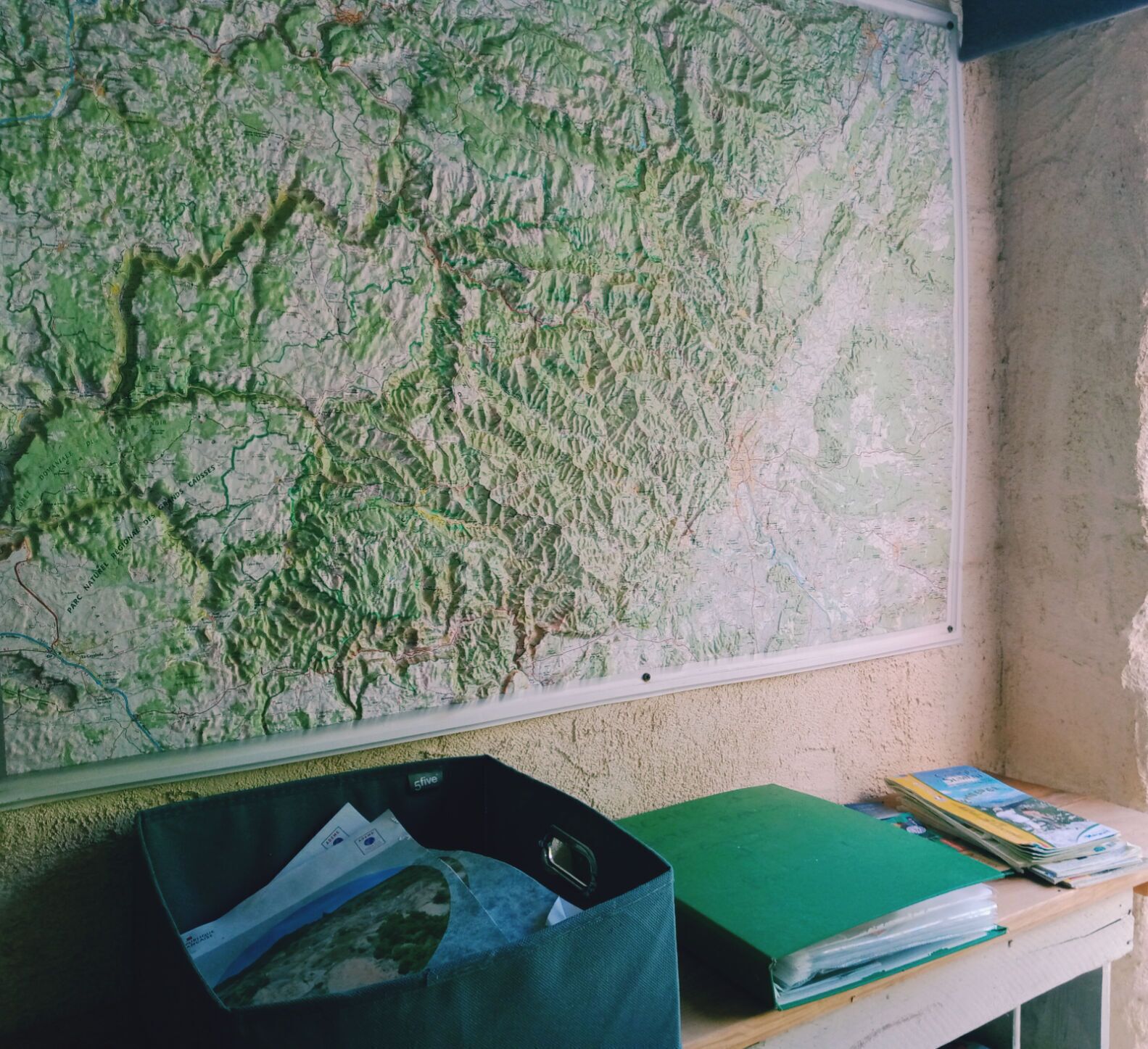 Une carte accrochée au mur, un classeur vert et une pile de livres sur un bureau.