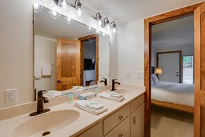 Une salle de bain avec deux vasques, un grand miroir et une porte en bois donnant sur une chambre.