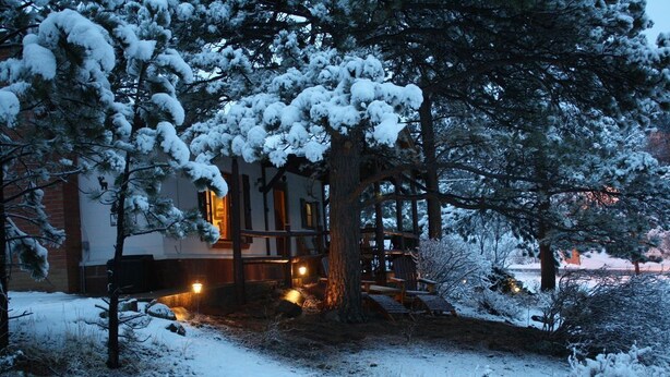 A snow-covered lodge with a wooden exterior, a deck, and a lit interior. - Close to Downtown Estes Park, RMNP, Hot Tub, Activity Shed! Far View Cabin (Estes Park)