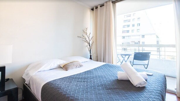 A hotel room with a bed, a small table with a chair, a vase with branches, and a view of buildings through the window. - Apoquindo Green (Santiago)