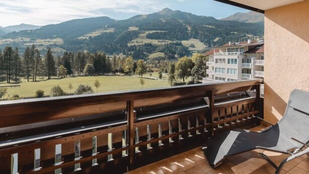 A balcony with a wooden railing and a single lounge chair, overlooking a mountainous landscape with buildings in the distance.