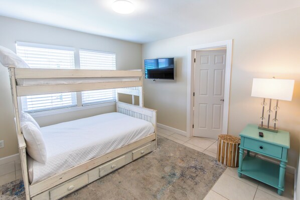 Quarto com cama de solteiro, televisão na parede, porta e pequena mesa de cabeceira com abajur.