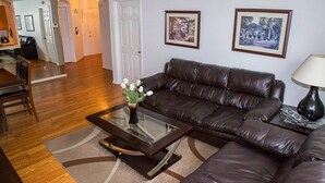 A living room with a brown leather sofa, a glass coffee table with a vase of flowers, and two framed pictures on the wall.