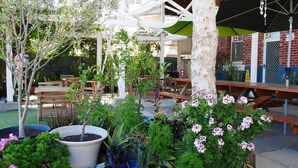 An outdoor patio area with wooden tables and chairs, surrounded by potted plants and flowering shrubs.