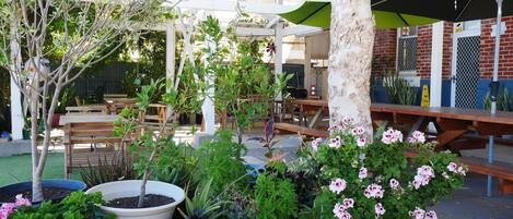 Un espace extérieur aménagé en terrasse, avec des tables et des chaises en bois, entouré de plantes en pot et d’arbustes en fleurs.