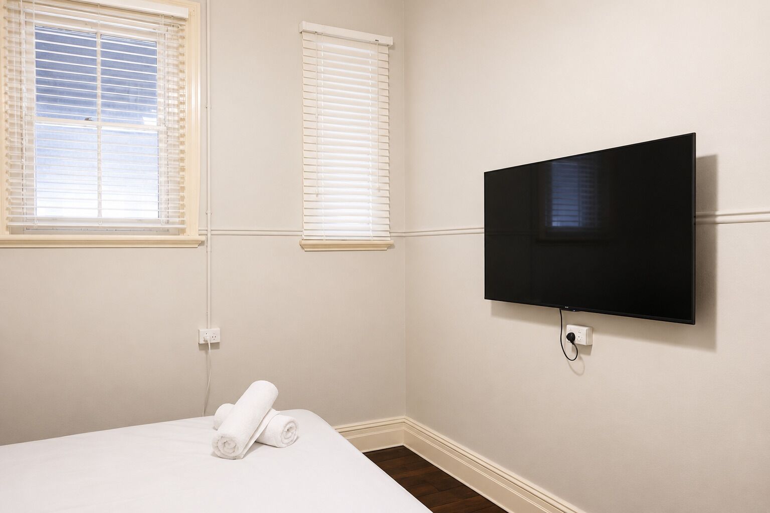 Habitació d'hotel amb un llit, dues finestres amb persianes i una televisió penjada.