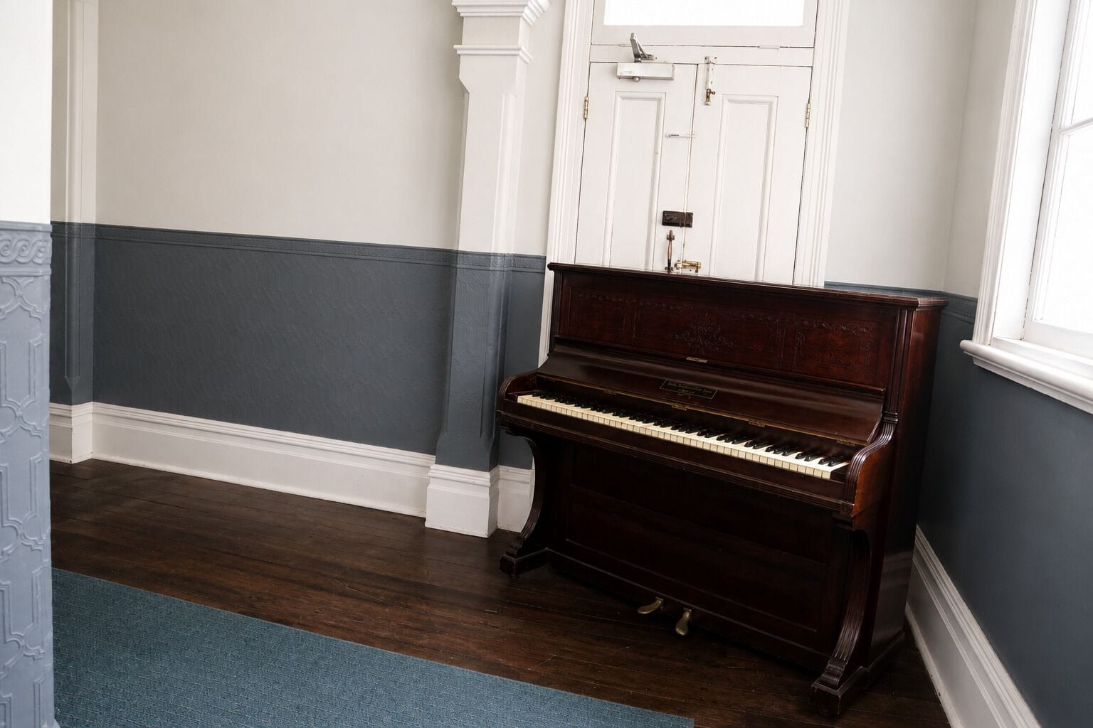 A piano in a room with a dark wooden floor, white and dark blue walls, and a white door in the background.