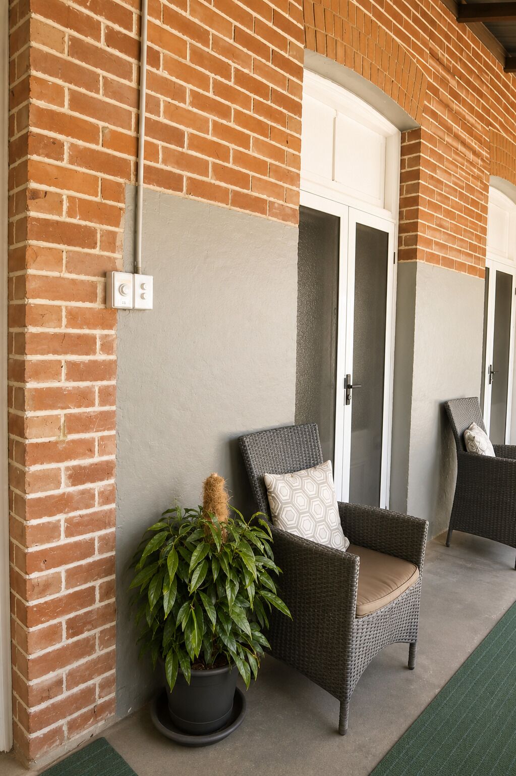 Pared exterior de ladrillo con una puerta blanca, una planta en maceta y dos sillas de mimbre con cojines.