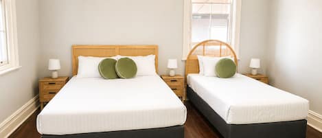 A double room with two beds, each with a wooden headboard, white bedding, and green pillows. There are bedside tables with lamps, a green rug on the floor, and a window with a blind.