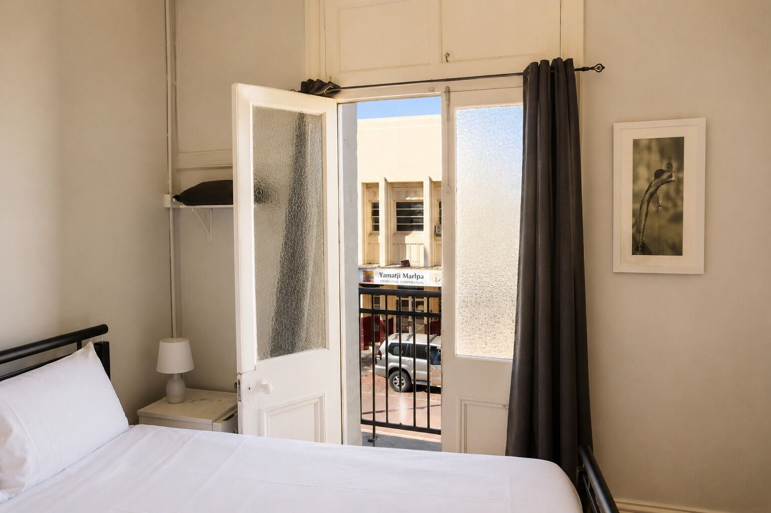 A hotel room with a bed, a nightstand, a lamp, a window with a view of the street, and a door leading to an outdoor area.