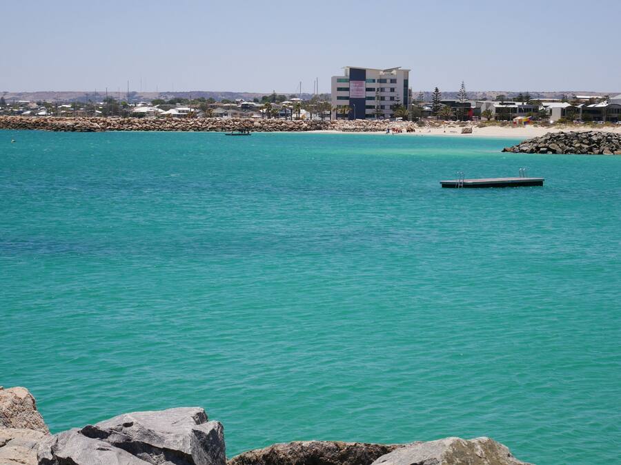 Vista costera con un mar turquesa y transparente, una costa rocosa y un edificio de varios pisos al fondo.