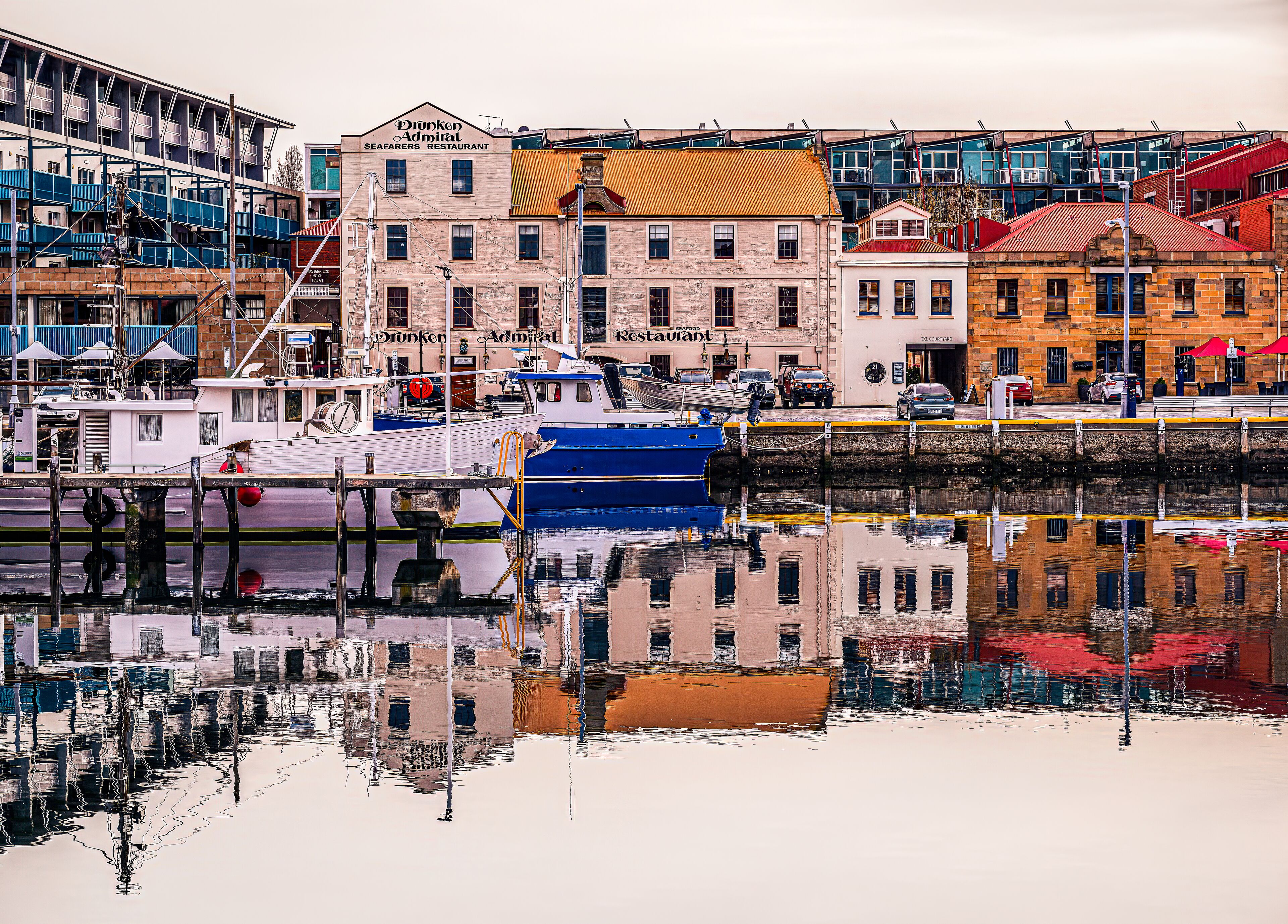 A waterfront with a blue boat docked at a pier, a building with 'Drunken Admiral' signage, and a reflection in the water.