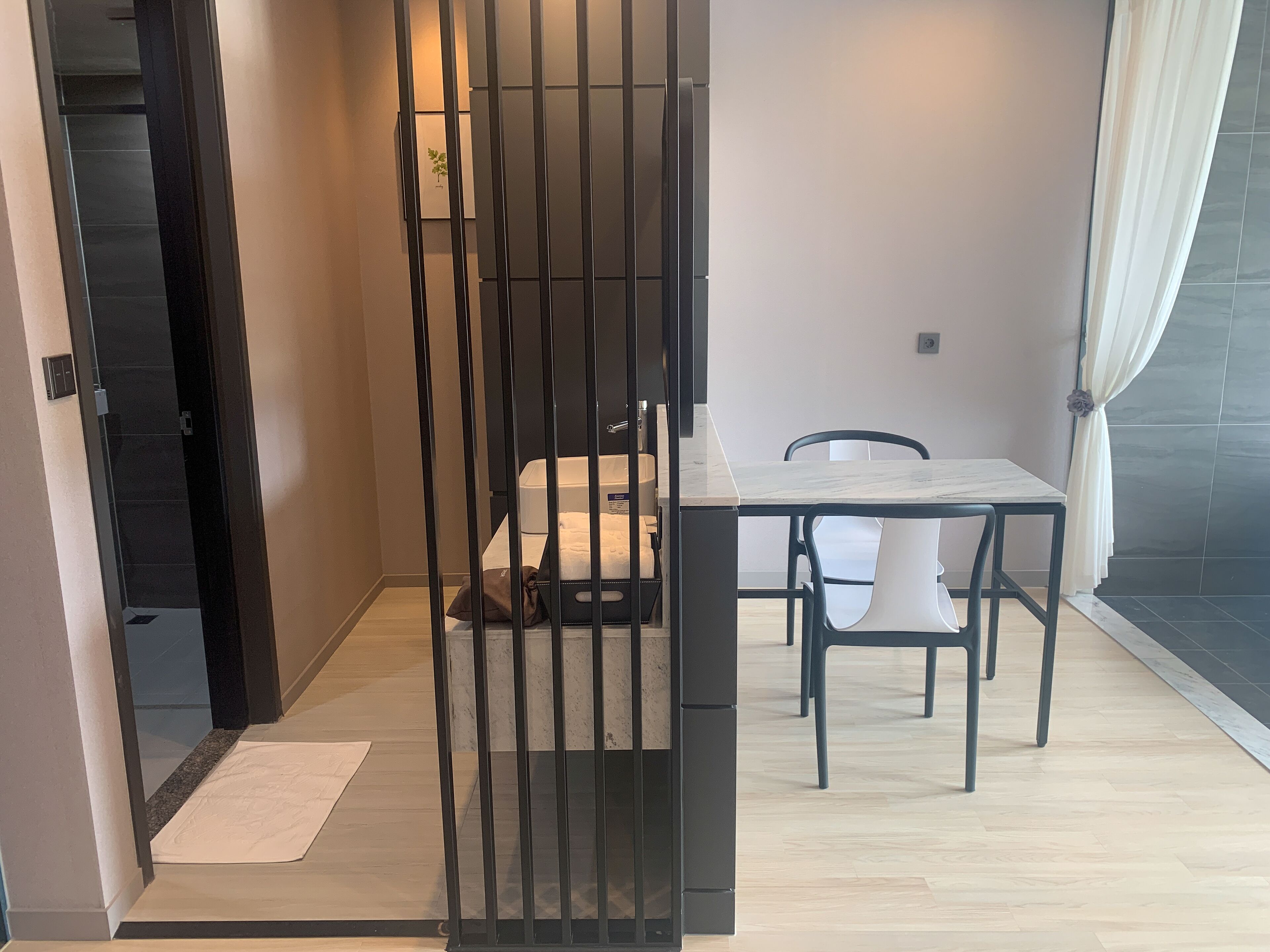 Una habitación moderna y funcional, con piso de madera, una pequeña mesa con dos sillas, una cama con colchón blanco y un separador de metal negro.