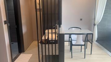 Una habitación moderna y funcional, con piso de madera, una pequeña mesa con dos sillas, una cama con colchón blanco y un separador de metal negro.