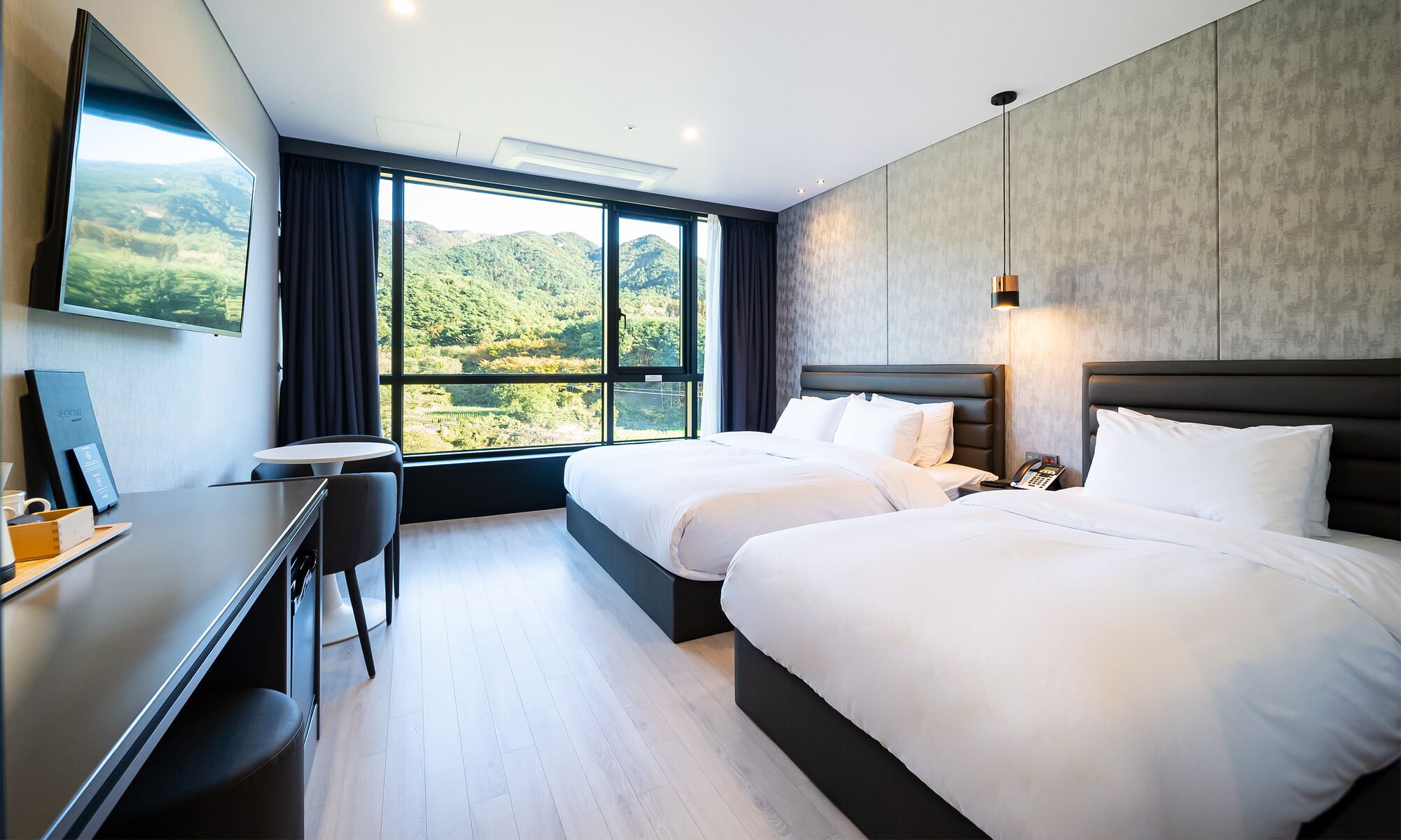 A hotel room with two beds, a desk, and a large window offering a scenic view.